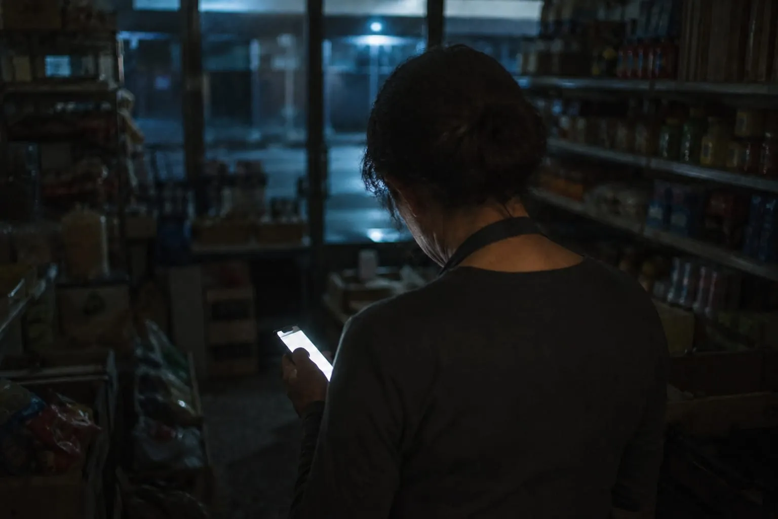 Un commerçant seul le soir dans sa boutique fermée, tête baissée, le visage éclairé par l’écran de son téléphone.